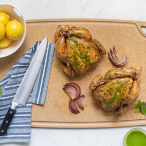 Sage 24" x 16" Carving Board With Non-Skid Feet with lemons, knife and two roasted chickens.