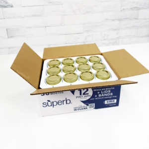 Open box of 12 pint-size canning jars with gold lids from Superb, neatly arranged in a white storage tray on a countertop with a white tile background.