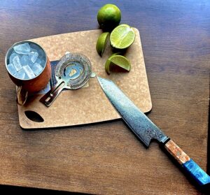 Sage cutting board on dark surface displaying knife, cup with ice, halved and quartered limes, and strainer spread out across cutting board. 