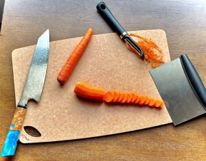 Sage cutting board on dark surface with knife, carrots, and peeler on top. 